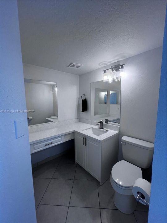 Bathroom featuring an L-shaped quartz countertop, white cabinetry with silver-tone hardware, a rectangular undermount sink, ceramic tile flooring, and a three-light fixture