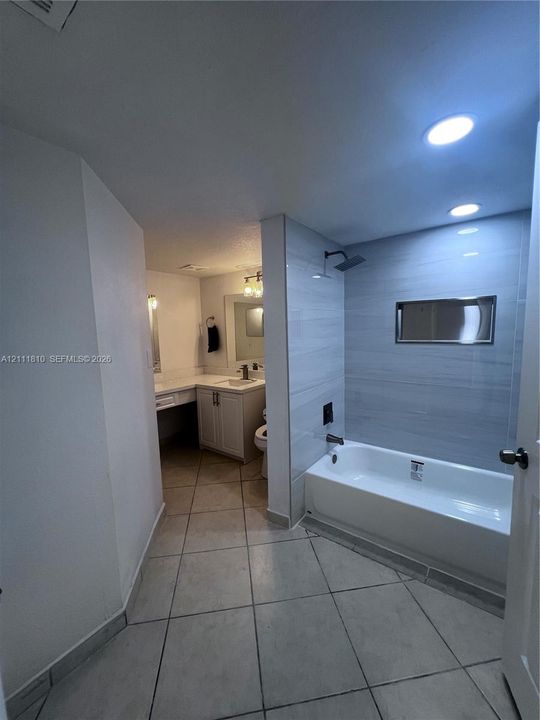 Bathroom featuring a soaking tub with a tiled surround, recessed wall niche, matte black rainfall shower fixture, and vanity cabinetry with a stone countertop