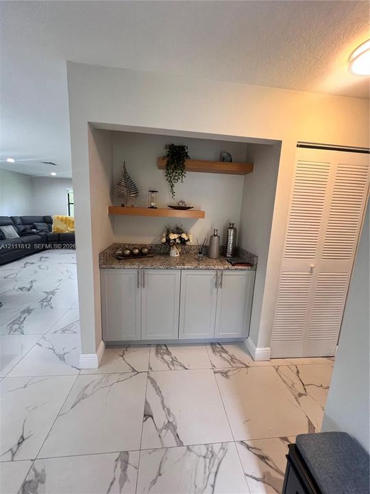 Built-in cabinetry featuring light-toned shaker doors, stone-finish countertops, floating wood-finish shelving, and white louvered closet doors, set against large-format tile flooring