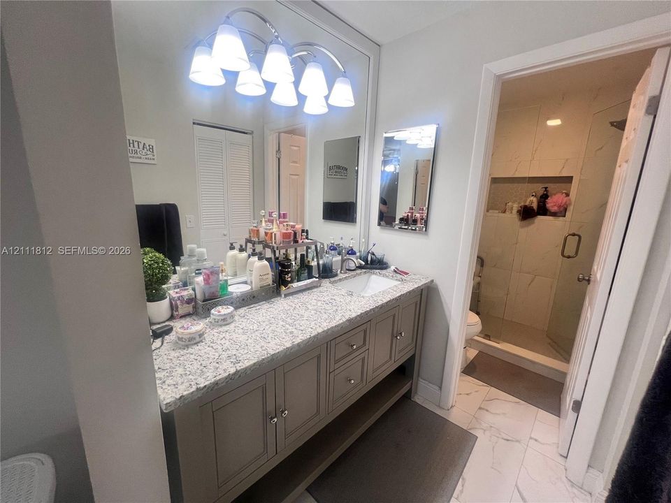 Vanity featuring a stone-composite countertop, undermount sink, shaker-style cabinetry, and five-light vanity fixture, complete with a walk-in shower featuring large-format tile walls and an integrated recessed niche
