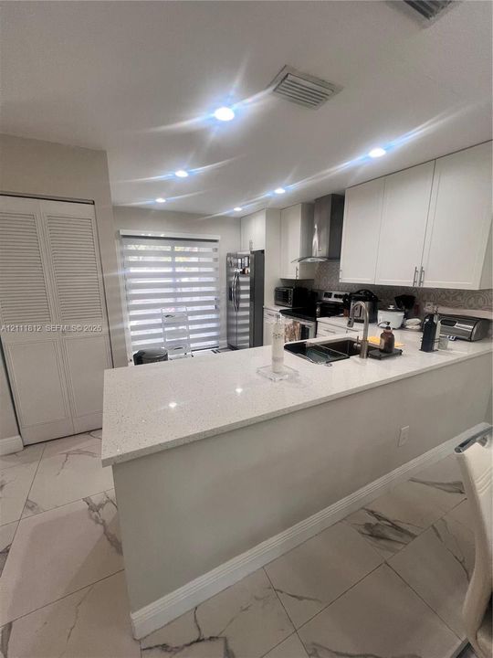 Kitchen featuring white Shaker-style cabinetry, a quartz-surfaced peninsula, large-format tile flooring, a stainless steel range hood, and recessed lighting