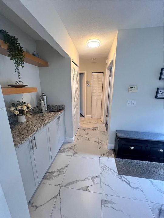 Hallway featuring large-format veined tile flooring, built-in lower cabinetry with stone-finish countertops, floating wood shelves, and louvered-door closets