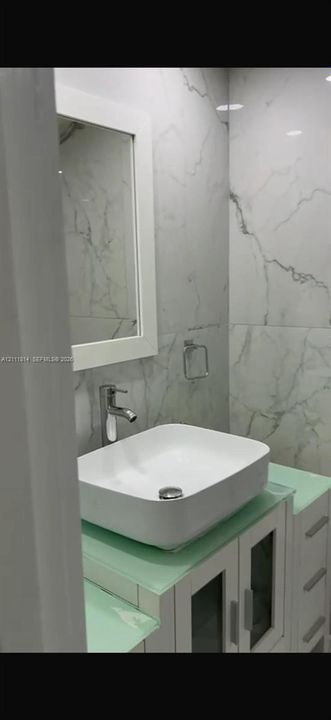 Bathroom featuring a vessel sink with a single-handle faucet, glass-top vanity with built-in cabinetry, framed mirror, and floor-to-ceiling veined tile wall surfacing