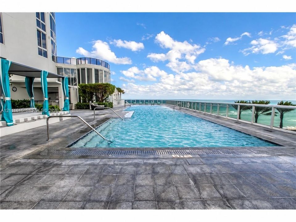 Infinity-edge swimming pool with metal handrails and stone tile decking, featuring a glass panel perimeter railing and oceanfront orientation