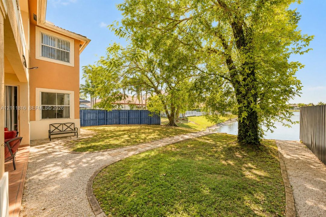 Waterfront backyard featuring a manicured lawn, stone-paved pathways, mature trees, and perimeter fencing