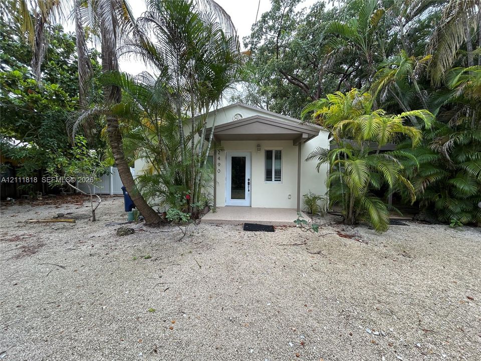 Exterior facade featuring a covered entryway, light-toned stucco siding, and a gravel ground surface surrounded by established palms and mature trees