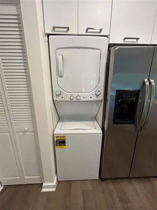 Stacked white laundry center featuring top-load washer and dryer, stainless steel side-by-side refrigerator, overhead white cabinetry with metallic pulls, and wood-finish flooring