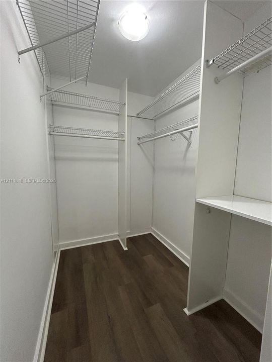 Walk-in storage space featuring wood-finish flooring, white built-in shelving partitions, overhead wire shelving, and a mounted ceiling light fixture