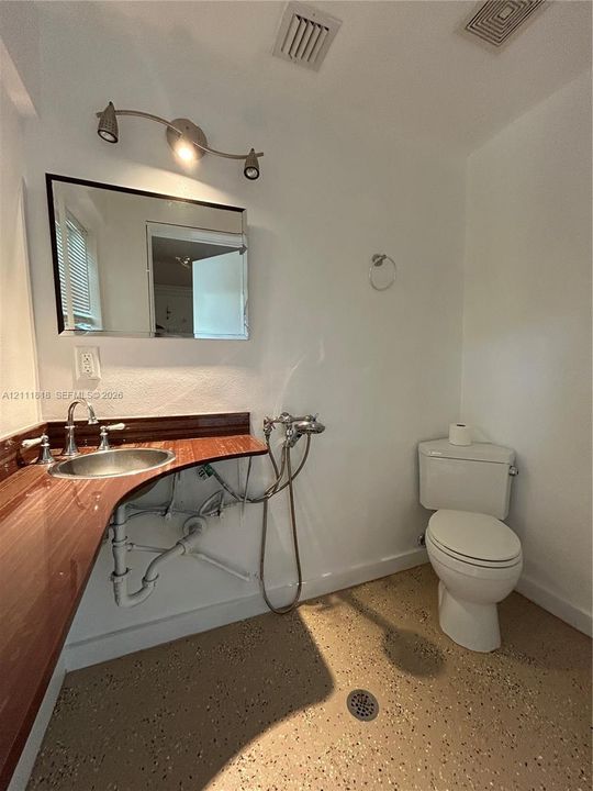 Bathroom featuring a solid-surface vanity countertop with an integrated metal sink, wall-mounted mirror, polished metal multi-bulb vanity light, and floor-level drain