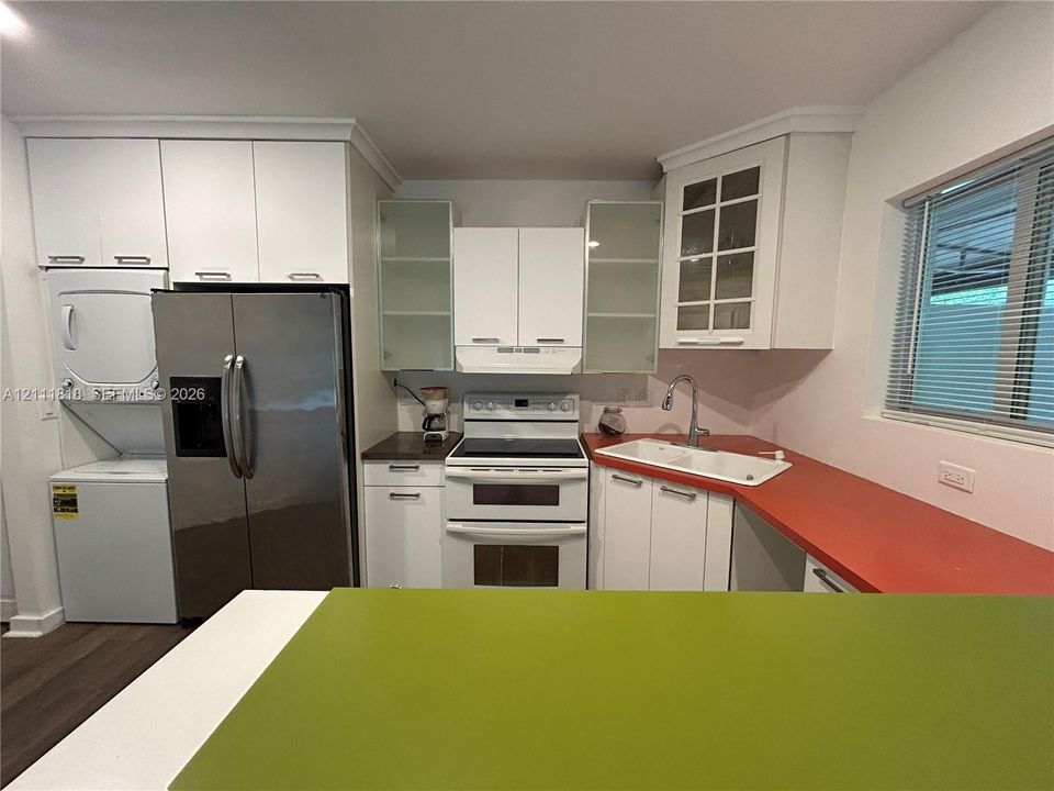 Kitchen featuring white cabinetry, glass-front upper cabinet doors, stainless steel refrigerator, white dual-basin sink, and stacked laundry unit
