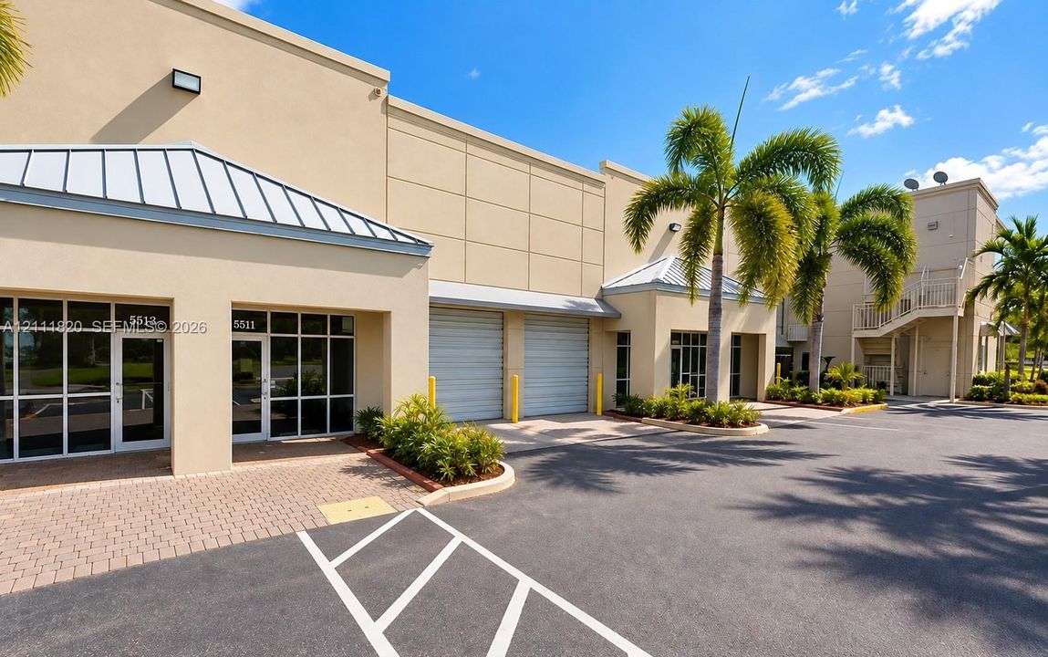 Commercial exterior featuring stucco facade, metal roll-up doors, multi-panel glass storefront entries, paved parking area, and palm tree landscaping