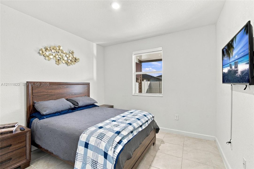 Neutral-toned bedroom featuring crisp white walls, large-format tile flooring, and a single-hung window with a view of a fenced exterior area