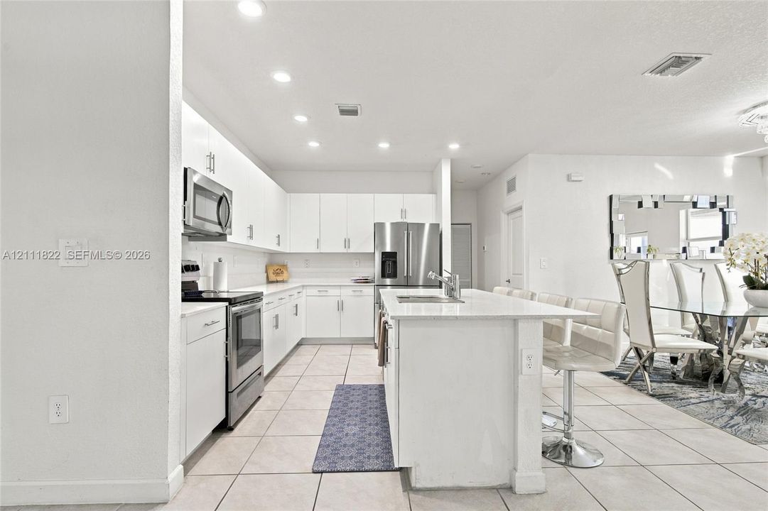 Kitchen featuring white shaker-style cabinetry, quartz countertops, stainless steel appliances, a central island with a built-in sink, and tile flooring