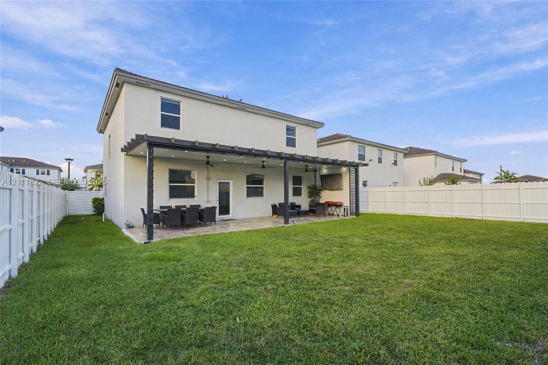 Rear exterior featuring a covered patio, pergola structure, exterior ceiling fans, and a perimeter privacy fence enclosing a lawn