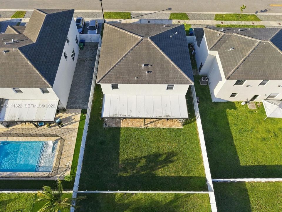 Aerial view of the rear exterior featuring a covered patio, paved hardscaping, a manicured lawn, and perimeter vinyl fencing