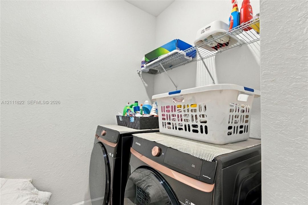 Laundry alcove featuring a pair of front-loading washer and dryer units with metallic copper-tone accents and overhead wire shelving