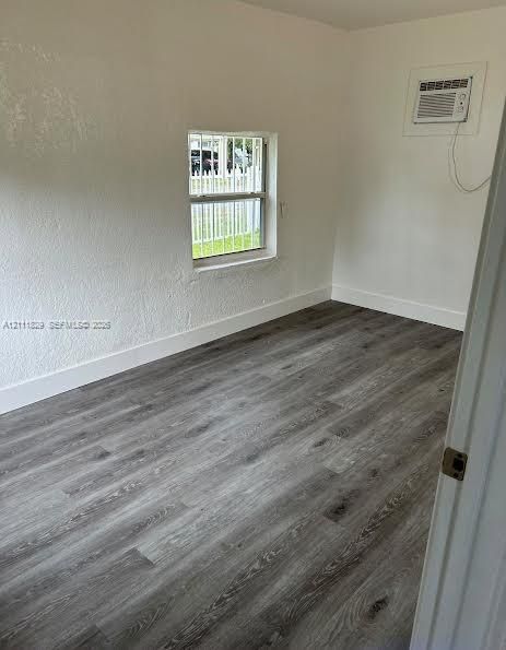 Interior space featuring wood-finish flooring, white walls, a single-pane window with security bars, and an integrated wall-mounted air conditioning unit