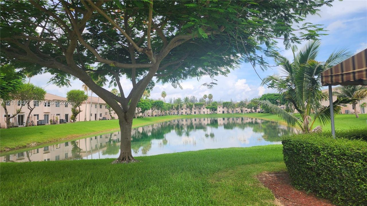 Waterfront exterior featuring a central pond, manicured lawn, mature shade trees, and groomed shrubbery