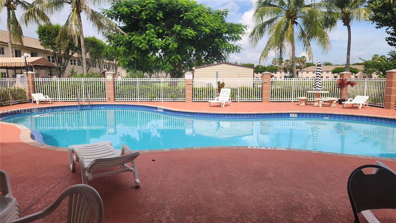 In-ground swimming pool featuring a curved design, masonry pool decking, and a perimeter white metal fence with masonry pillars