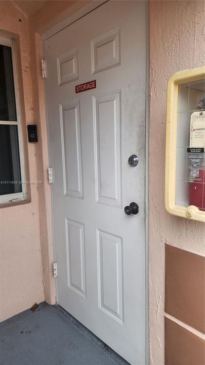 Six-panel white exterior storage door featuring a deadbolt lock and cylindrical knob, complemented by a wall-mounted glass-enclosed utility cabinet