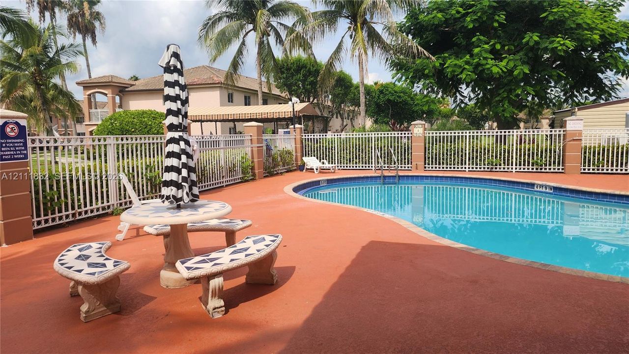 Curved swimming pool with tiled coping, surrounded by a paved deck and enclosed by white metal fencing