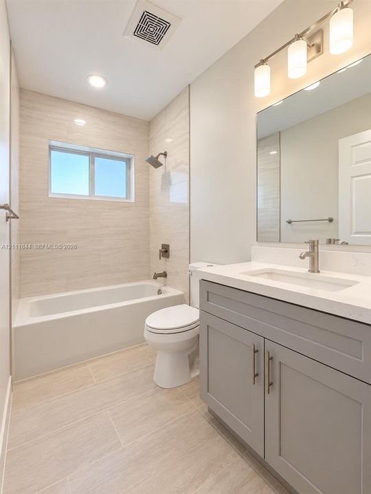 Bathroom featuring a gray vanity with a stone countertop, an integrated rectangular sink, neutral large-format wall and floor tile, and a bathtub with showerhead and mixer faucet