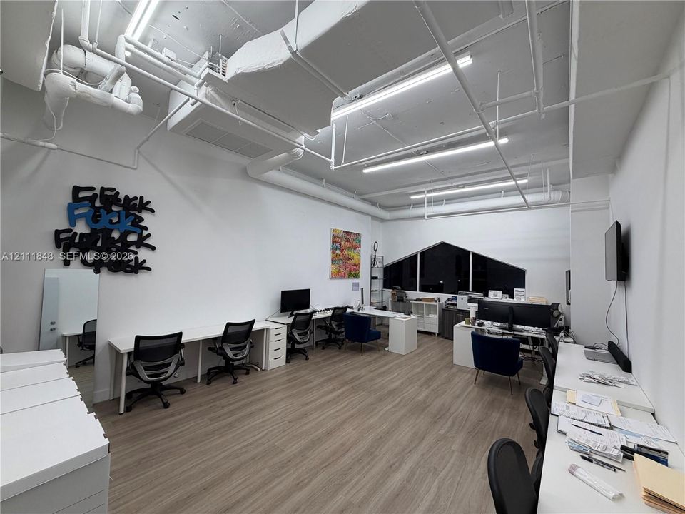 Industrial-style workspace featuring high exposed ceilings with linear lighting, wood-finish flooring, and a large custom-shaped window