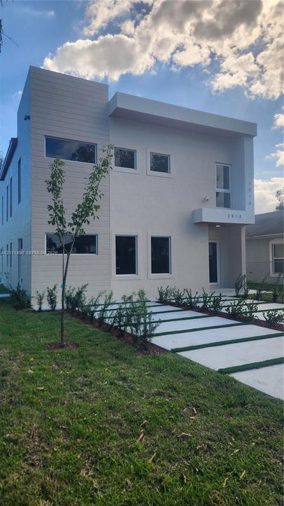 Modern two-story exterior featuring a horizontal siding facade, flat roofline, rectangular window configuration, and a concrete paver driveway with turf insets