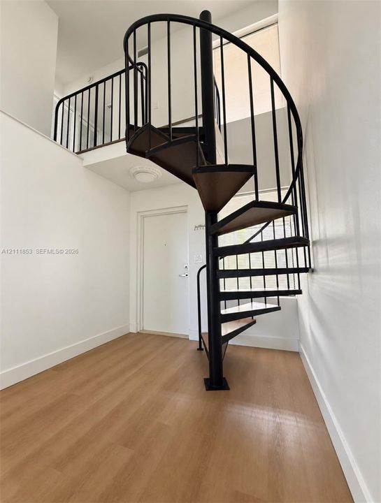 Spiral staircase featuring a black metal frame, wooden treads, and matching railing, complemented by wood-finish flooring and a mezzanine landing