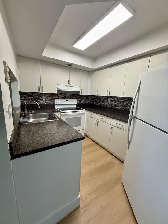 Kitchen featuring white shaker-style cabinetry, dark stone-finish countertops, multi-tonal tile backsplash, integrated stainless steel sink with gooseneck faucet, and wood-finish flooring