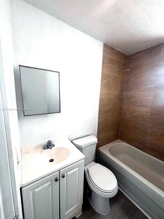 Bathroom featuring a wood-finish tile shower surround, a white vanity cabinet with an integrated sink, a rectangular wall-mounted mirror, and a standard porcelain toilet