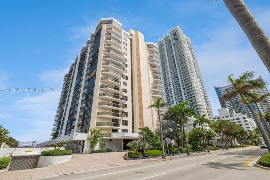 High-rise residential exterior featuring stacked private balconies, concrete construction, street-level covered parking access, and tropical landscaping