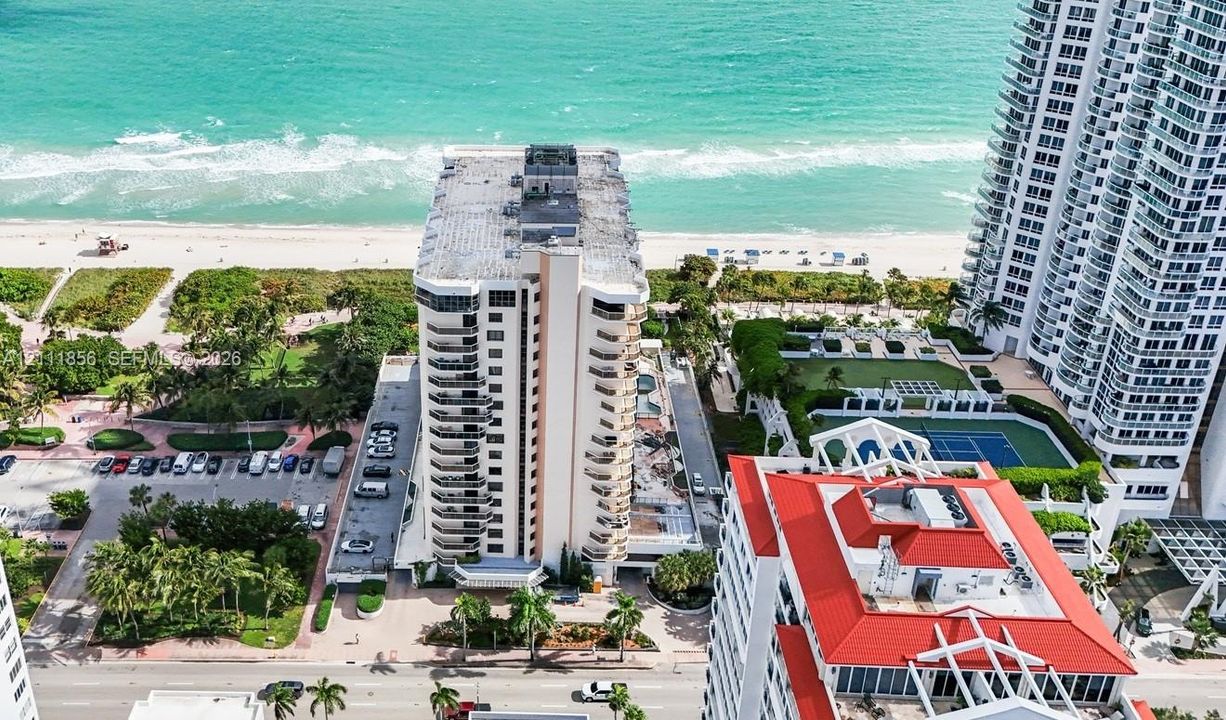 High-rise residential building featuring multiple private balconies, direct coastal frontage, paved surface parking, and shared tennis courts