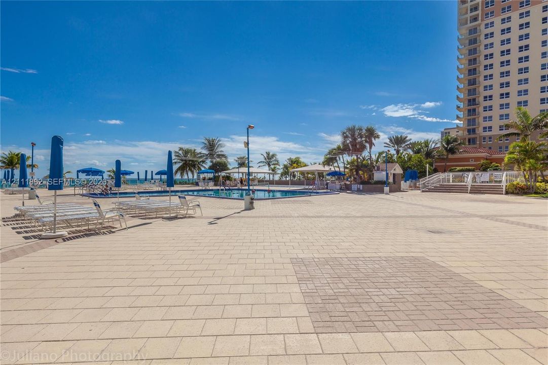 Outdoor swimming pool surrounded by a paved deck, shaded structures, and lush palm trees