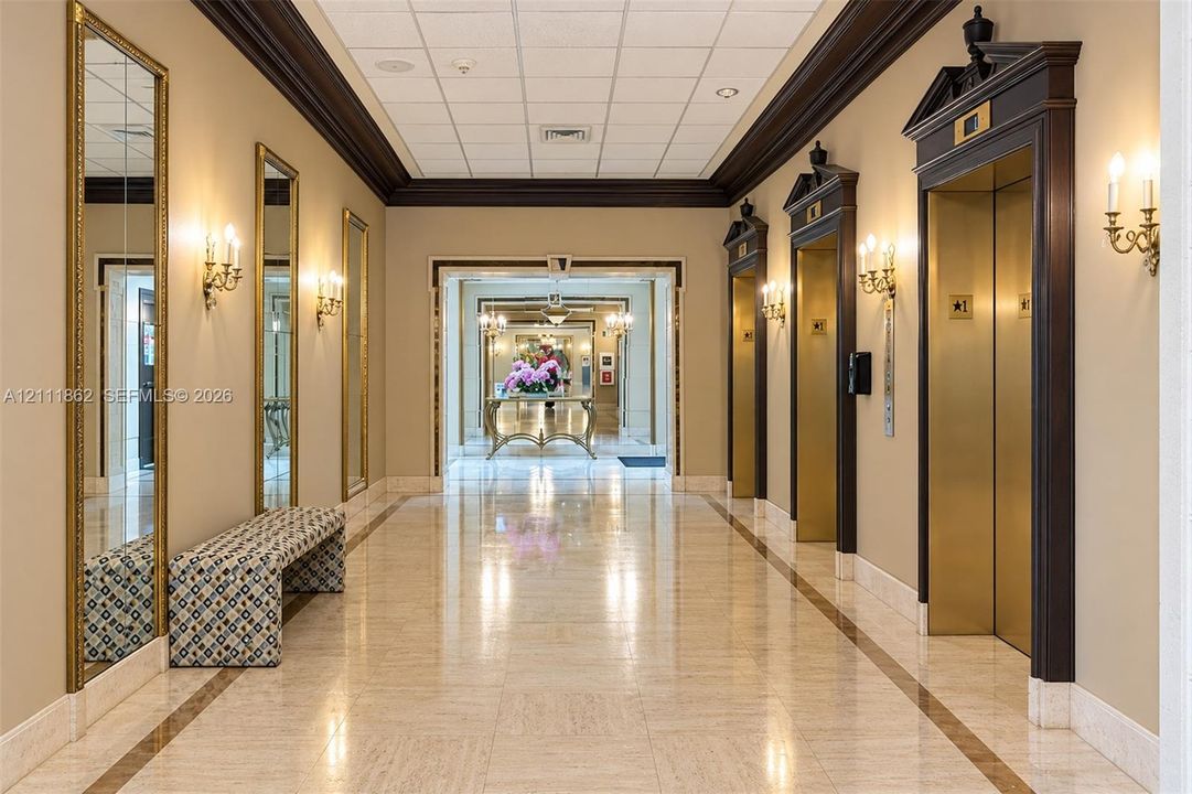 Formal corridor featuring polished stone flooring, custom dark-wood elevator surrounds with architectural pediments, integrated wall-mounted sconces, and full-length framed mirrors
