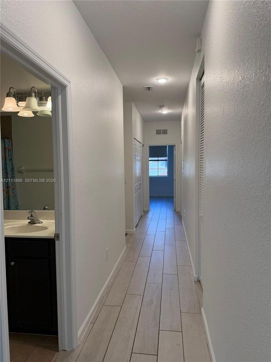 Hallway featuring wood-finish tile flooring, recessed lighting, and a bathroom containing a dark-toned vanity with a white countertop and multi-bulb light fixture