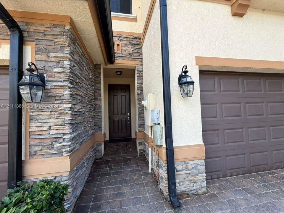 Exterior facade featuring multi-textured masonry stone veneer, smooth stucco siding with tan trim, a paver walkway, and carriage-style wall-mounted lanterns