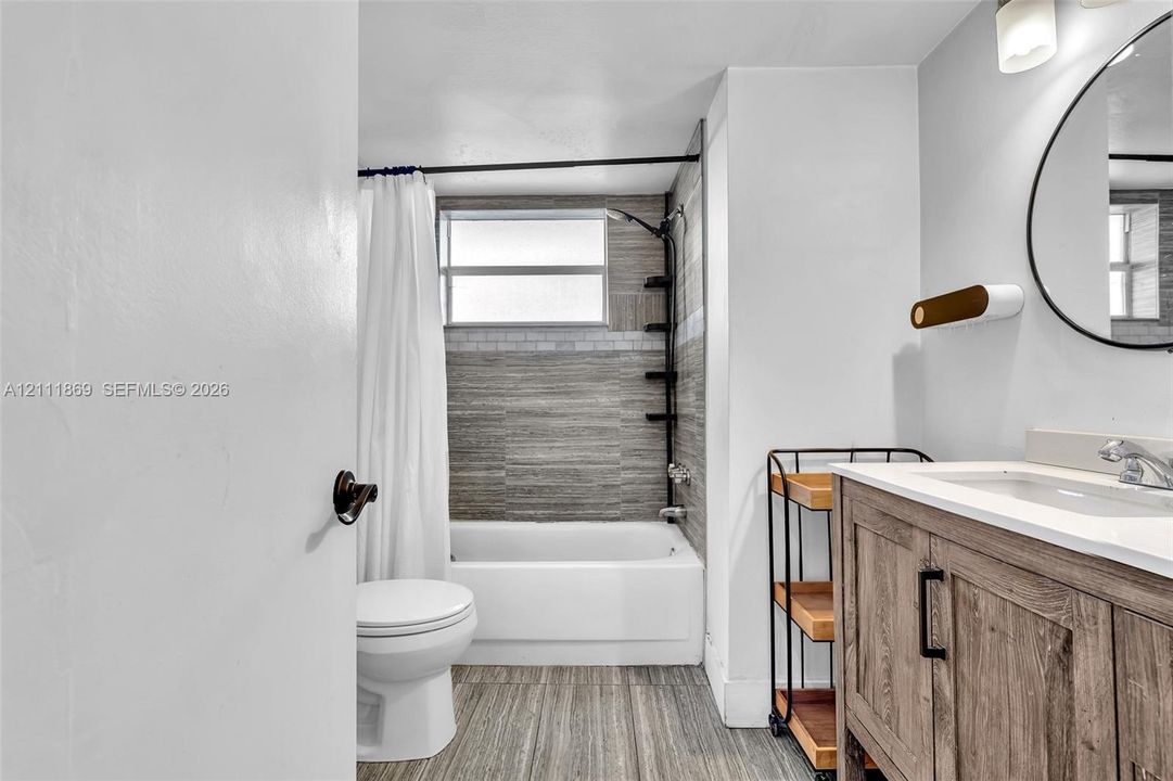 Bathroom featuring a wood-finish vanity with a stone countertop, circular wall-mounted mirror, soaking tub with tile surround, and wood-finish tile flooring