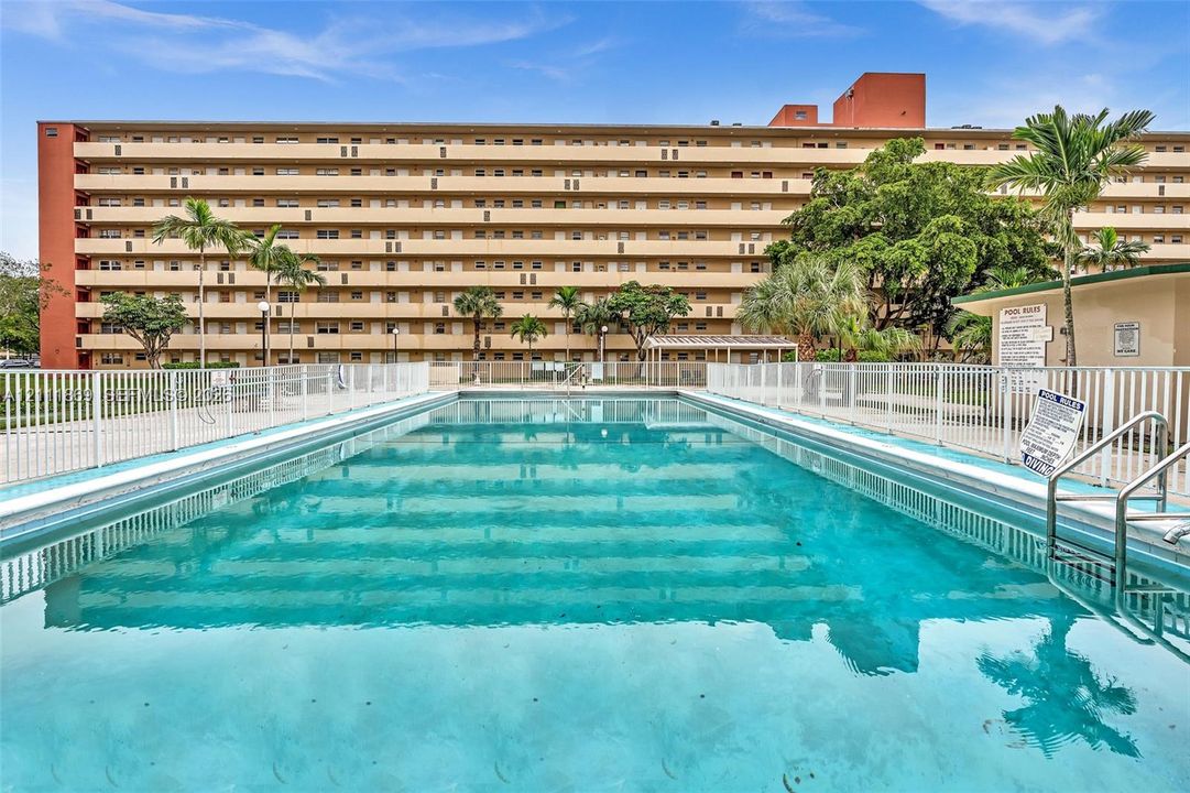 In-ground swimming pool featuring concrete steps and metal handrails, enclosed by white perimeter fencing with mature palm trees