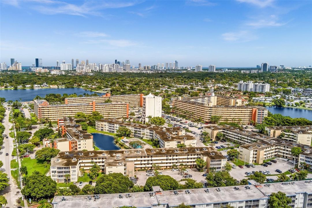 Aerial perspective of a multi-building residential complex featuring water retention areas, surface parking, and dense mature tree canopy with a distant metropolitan skyline backdrop