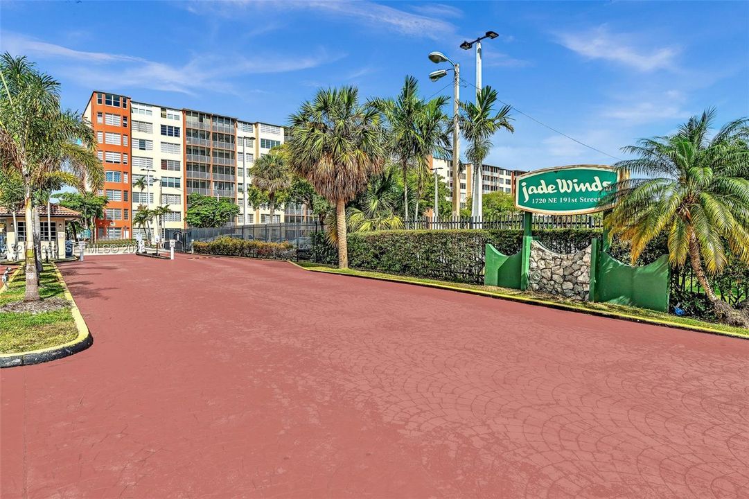 Gated entrance featuring stamped concrete paving, perimeter fencing, mature palm landscaping, and signage