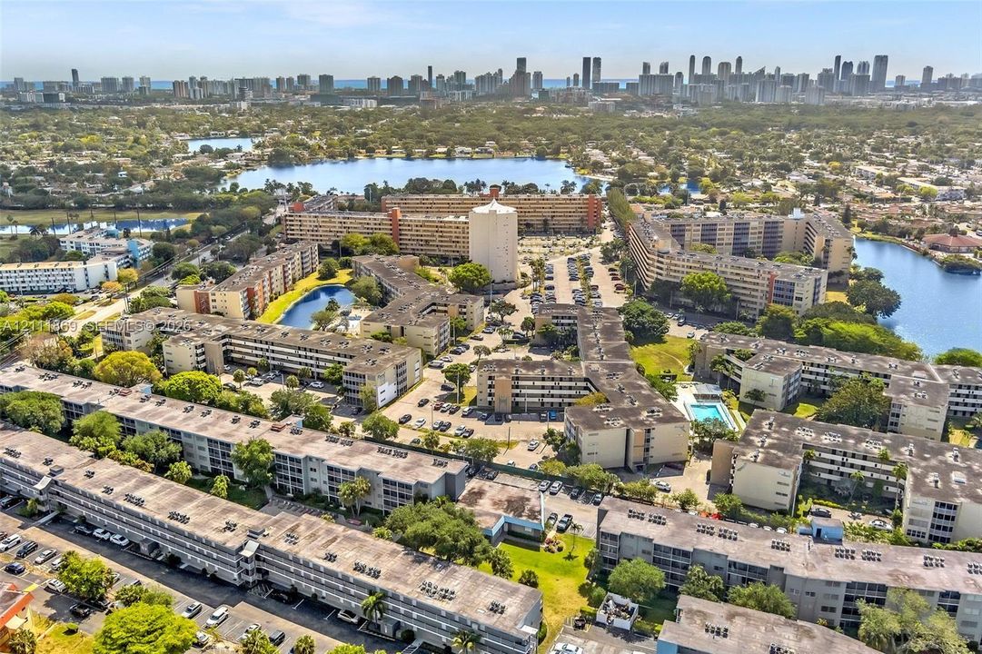 Aerial perspective of a multi-building residential complex featuring surface parking areas, landscaped grounds, multiple bodies of water, and an outdoor swimming pool with an urban skyline backdrop