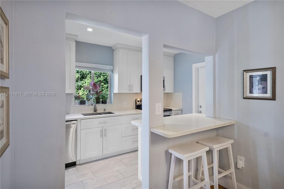 Kitchen featuring white shaker-style cabinetry, solid-surface countertops, dual-basin stainless steel sink, tile flooring, and a built-in pass-through breakfast bar