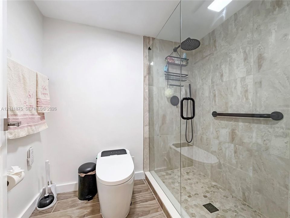 Walk-in shower with stone-tile surround, pebble flooring, fixed rainfall showerhead, and a frameless glass enclosure featuring matte black hardware