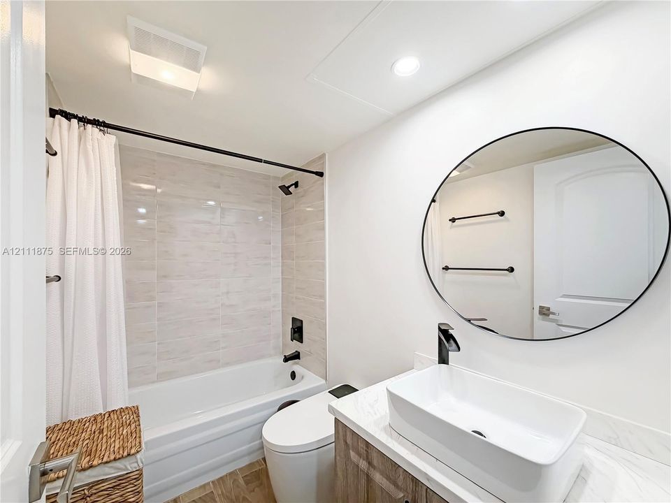 Bathroom featuring a floating vanity with a rectangular vessel sink and matte black hardware, stone-patterned countertop, circular wall-mounted mirror, and tile-surround shower and tub combination