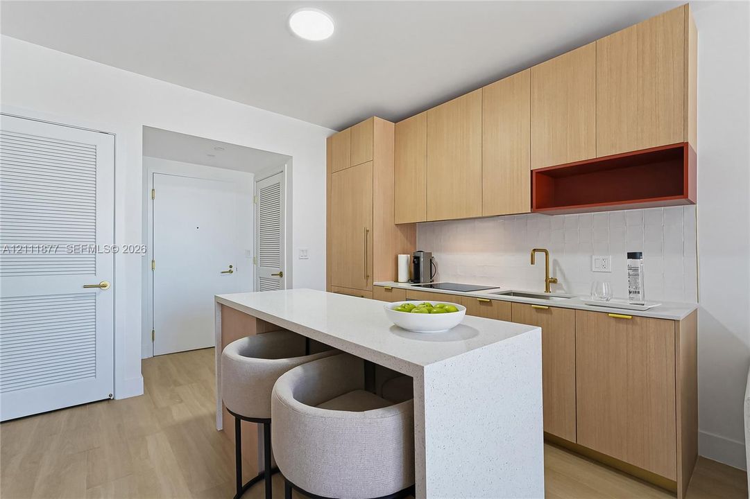 Kitchen featuring light-tone wood cabinetry, a solid-surface waterfall edge island, a contrasting open shelving unit, a brass-finish faucet, and white textured backsplash tile