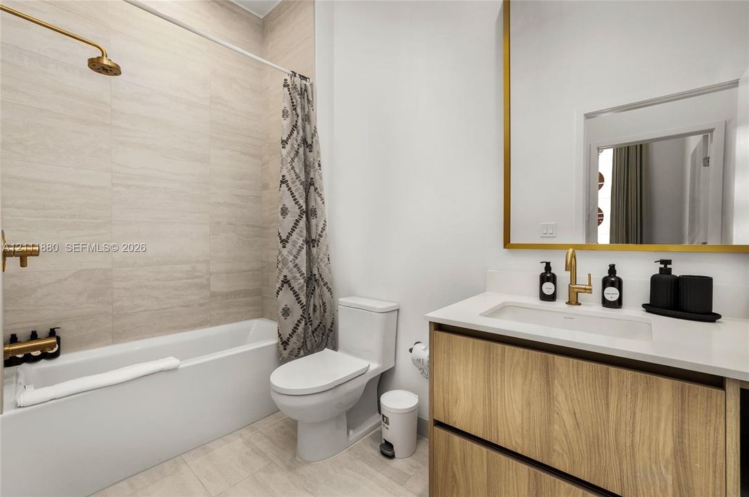 Bathroom featuring a wood-finish vanity with a stone countertop, a large rectangular mirror with a gold-toned frame, wall-mounted gold-toned plumbing fixtures, and floor-to-ceiling tile surrounding the bathtub