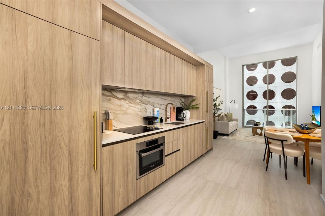 Contemporary kitchen featuring light wood-finish cabinetry, integrated appliances, a stone-pattern backsplash, solid surface countertops, and brushed gold-tone hardware