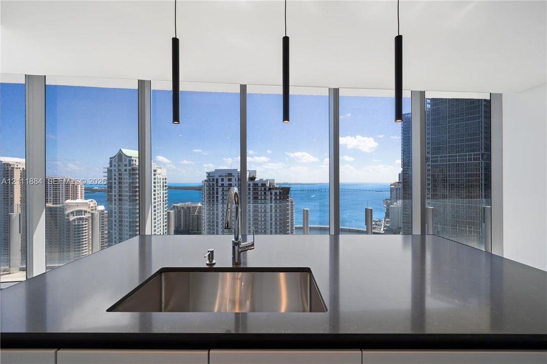 Kitchen island featuring dark solid-surface countertops, an undermount stainless steel sink with a high-arch faucet, floor-to-ceiling windows, and matte black cylindrical pendant lighting