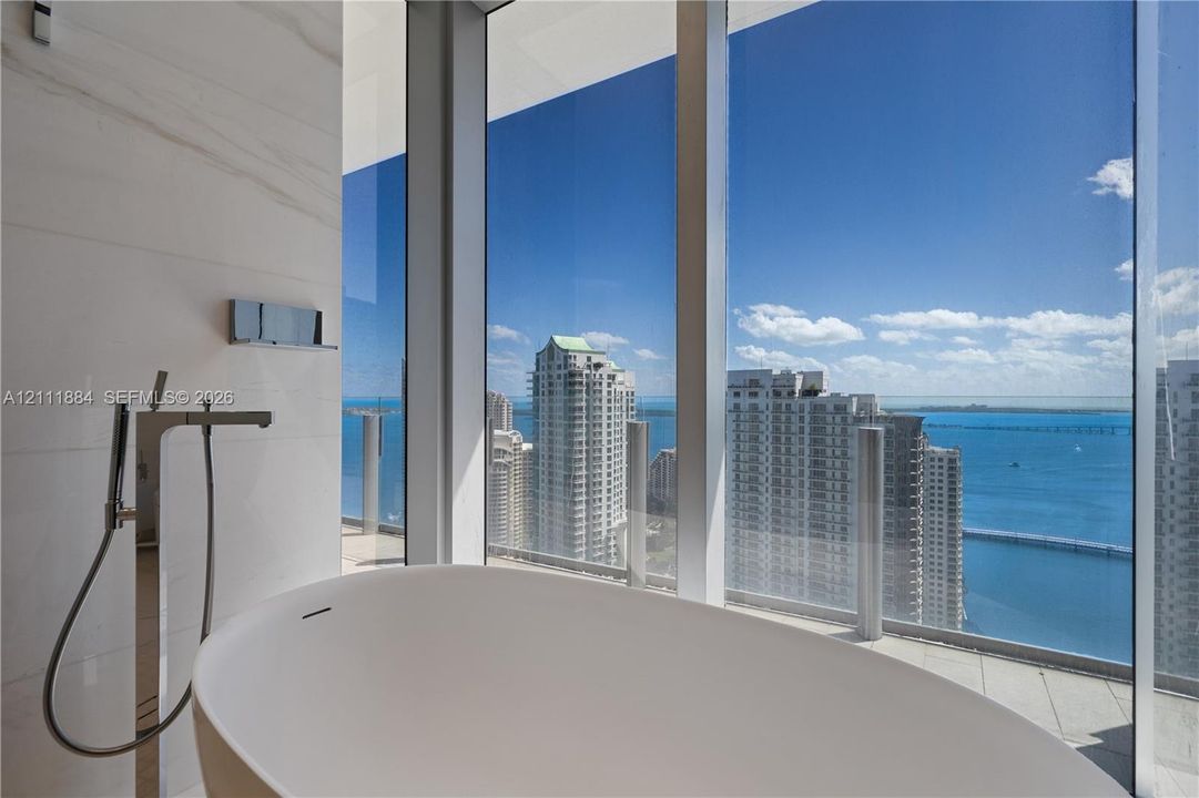 Floor-to-ceiling windows featuring panoramic water and city scenery, a white freestanding soaking tub, and wall-mounted metallic fixtures with a handheld shower wand